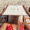 Person wearing a red bikini sitting on a beach chair with sun visor with a BLYND sign in front of their face.